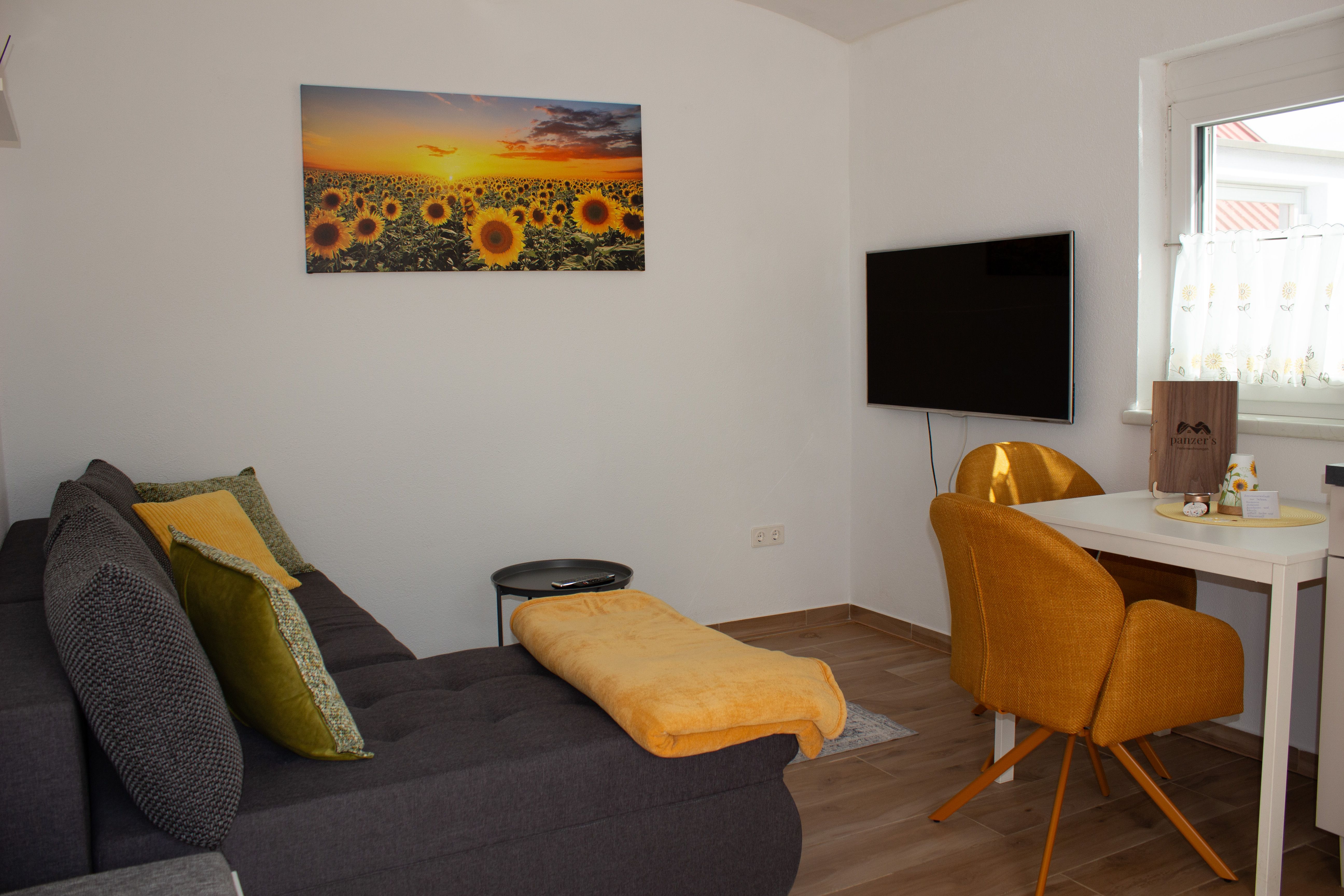 Living room of an apartment with a gray sofa, yellow cushions, a picture of sunflowers and a TV on the wall.