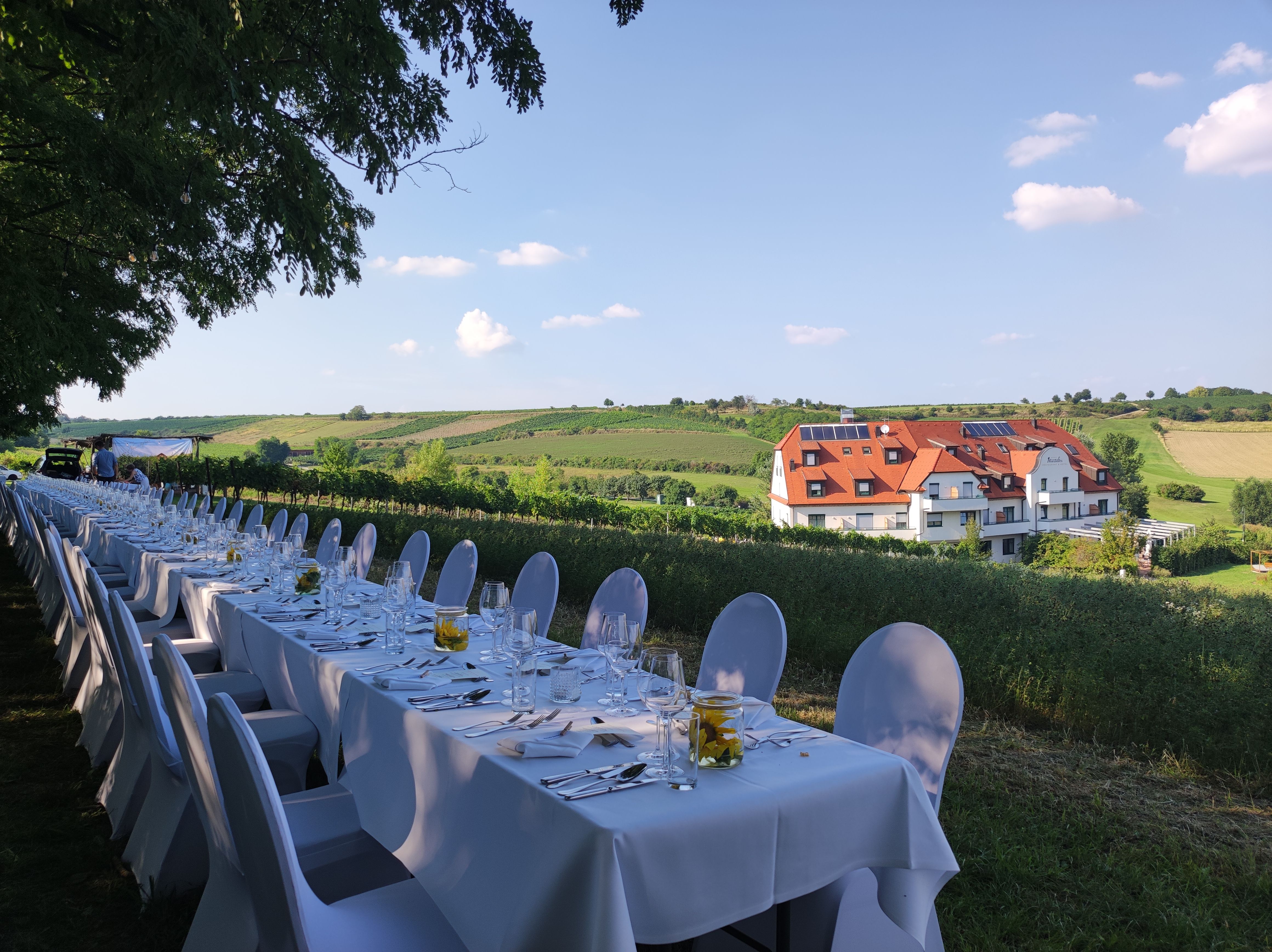 Lange Tafel im Freien mit weißer Tischdecke, gedeckt für ein Festessen, im Hintergrund ein Gebäude und grüne Felder.