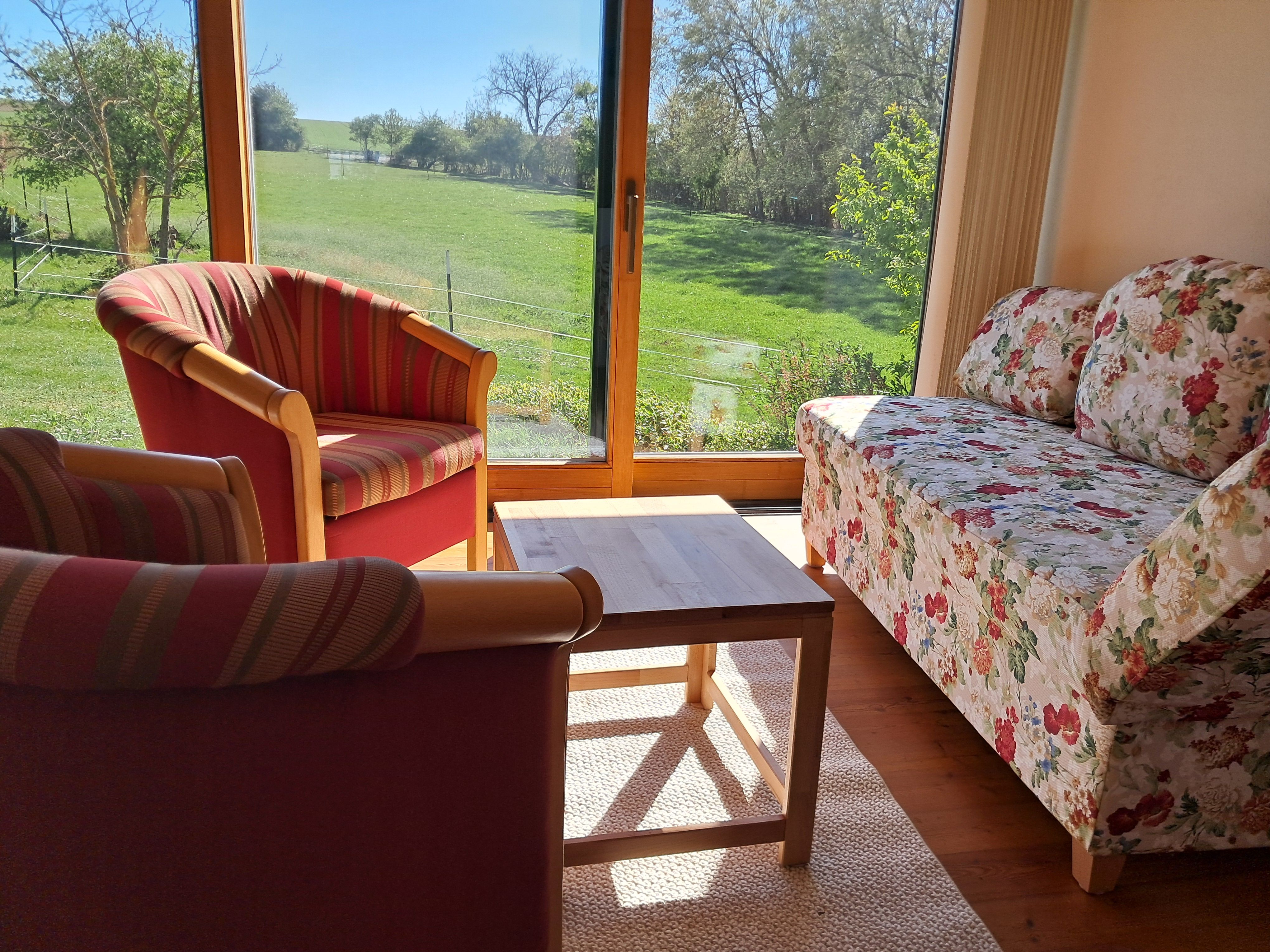 Wohnzimmer mit großen Fenstern, Blick auf grüne Wiese, zwei gestreifte Sessel, geblümtes Sofa und Holztisch.