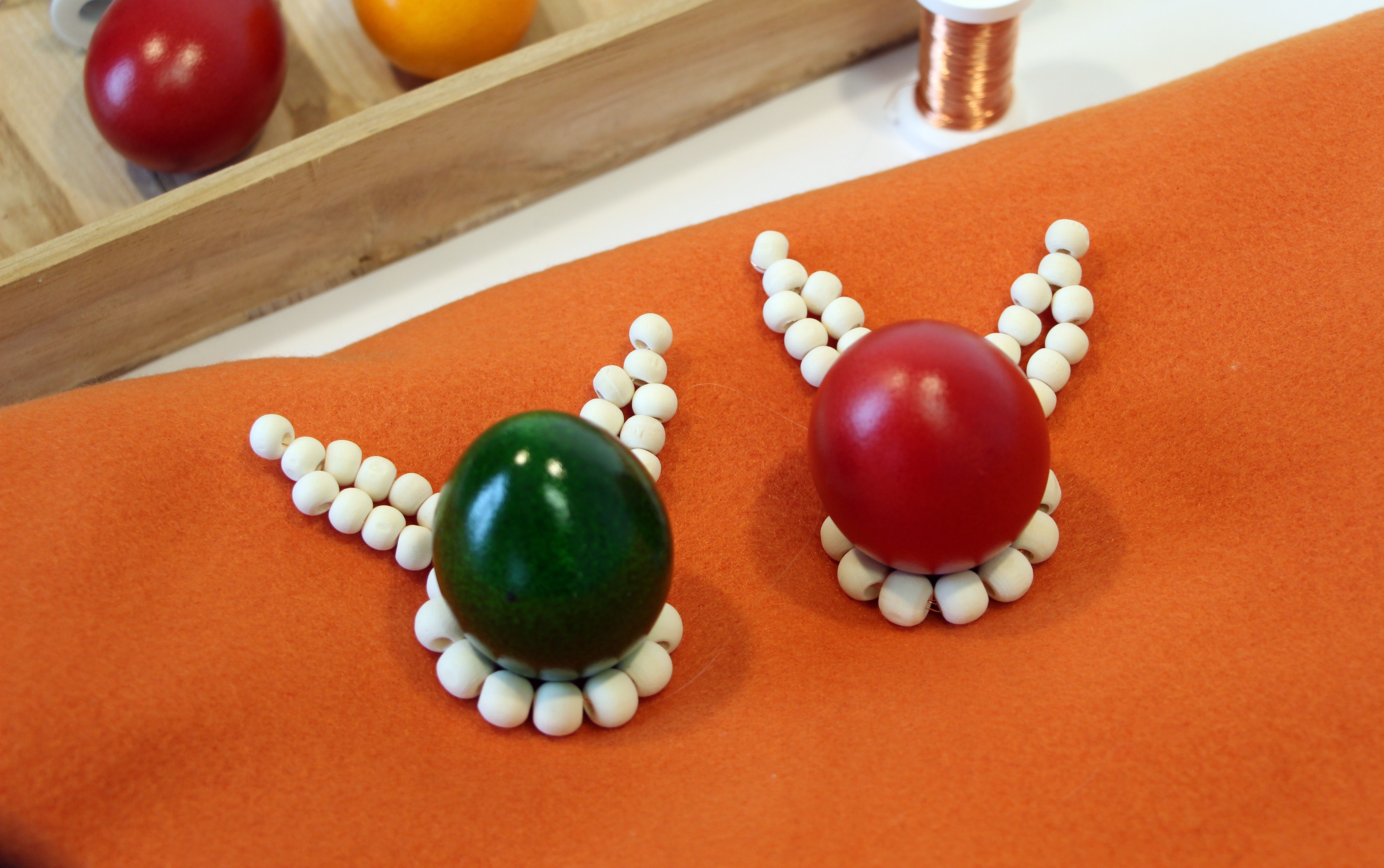 Zwei Eierbecher aus Holzperlen mit bunten Ostereiern sind auf einem Tisch platziert, im Hintergrund sieht man Eier und Bastelutensilien.