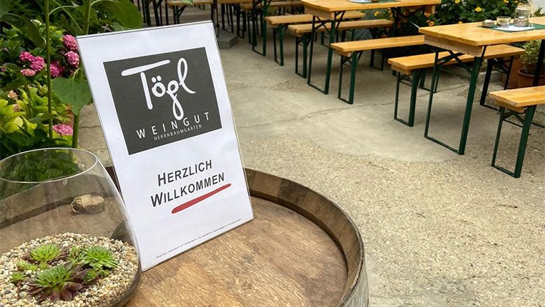 Ein Schild mit der Aufschrift 'Weingut Tögl' und 'Herzlich Willkommen' steht auf einem Weinfass. Im Hintergrund sind Biertische und Pflanzen zu sehen.