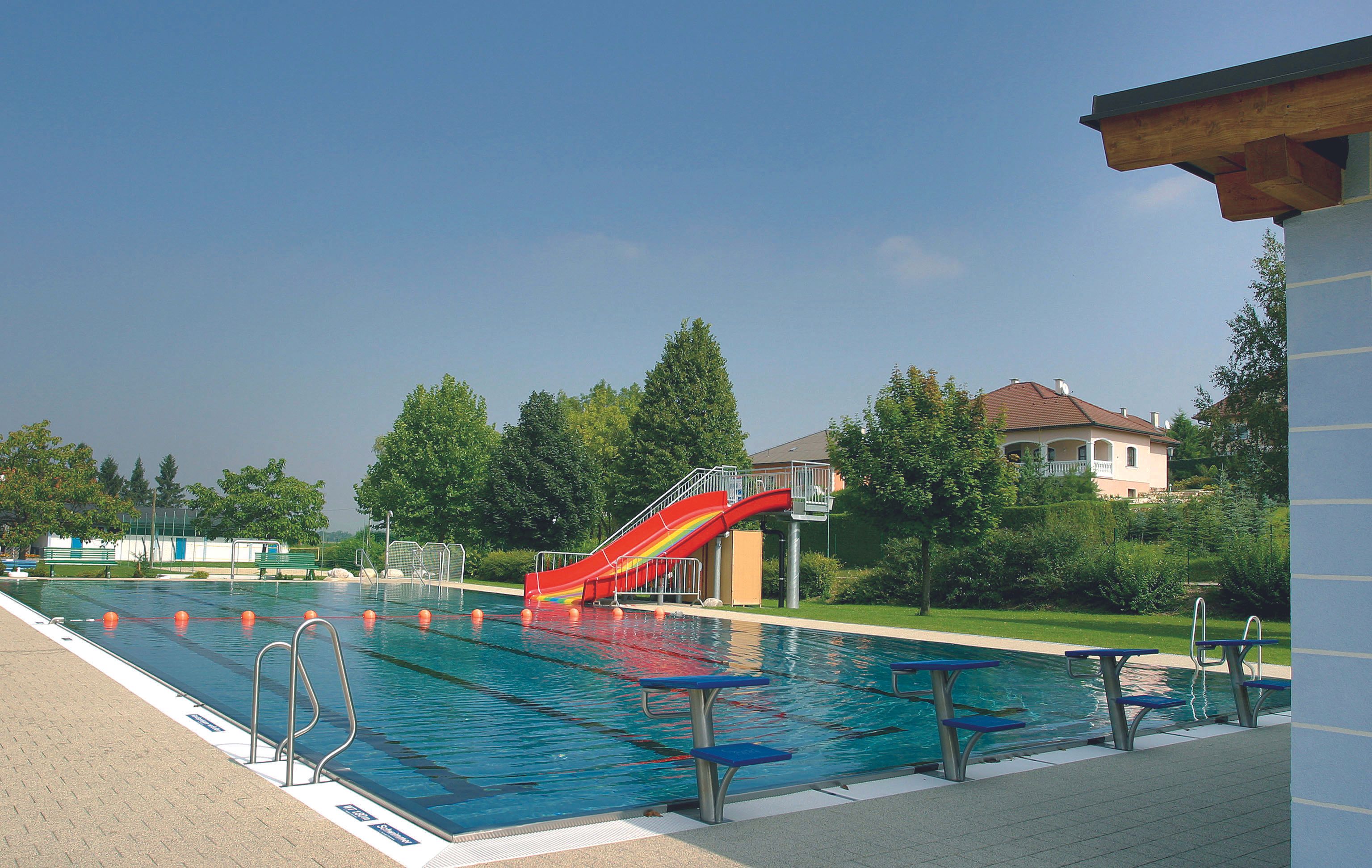 Ein Freibad mit einem Schwimmbecken, einer roten Rutsche und Startblöcken. Im Hintergrund sind Bäume und ein Wohnhaus zu sehen.
