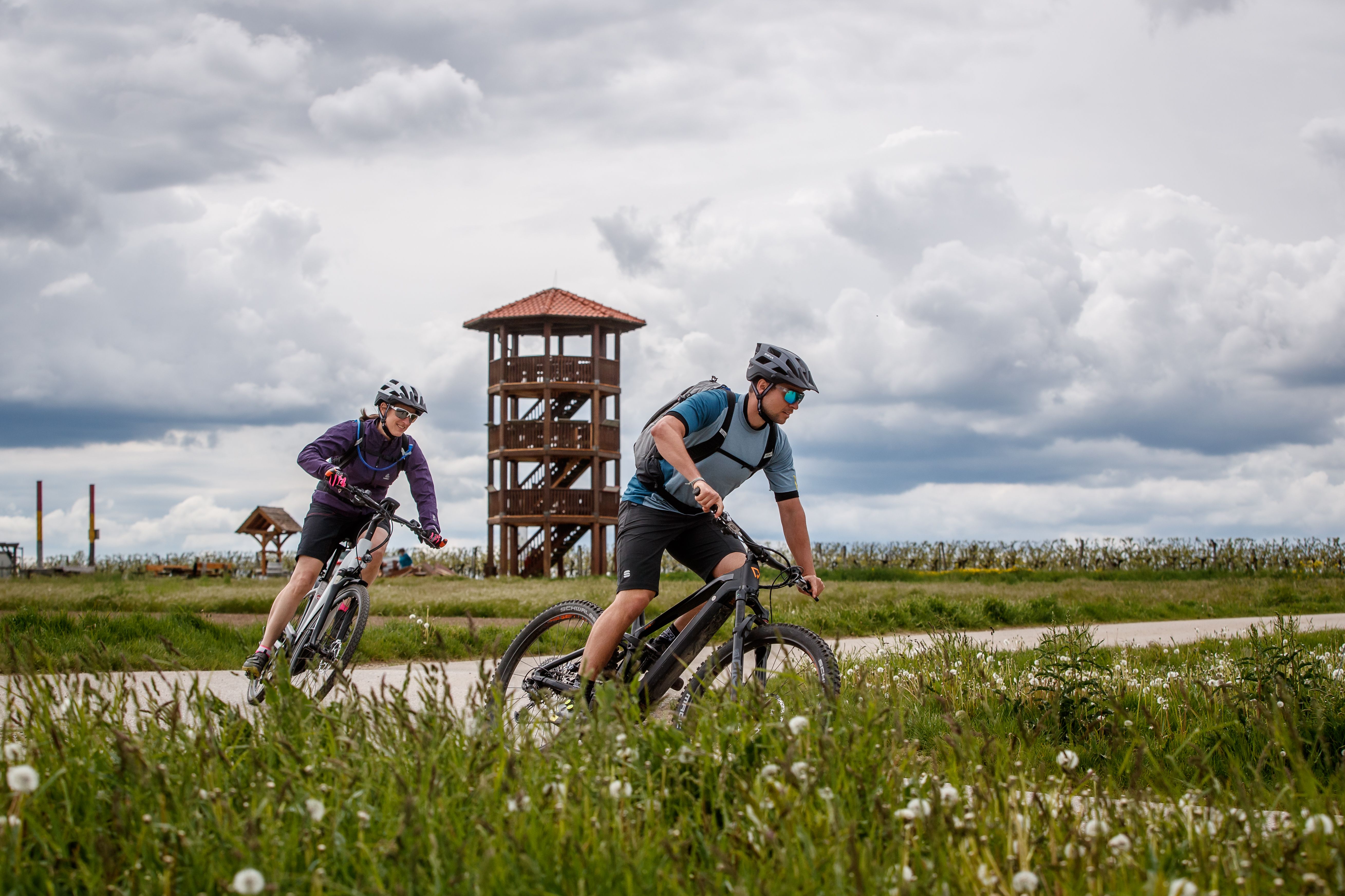 Two cyclists ride along a path in front of a wooden tower in a rural landscape.