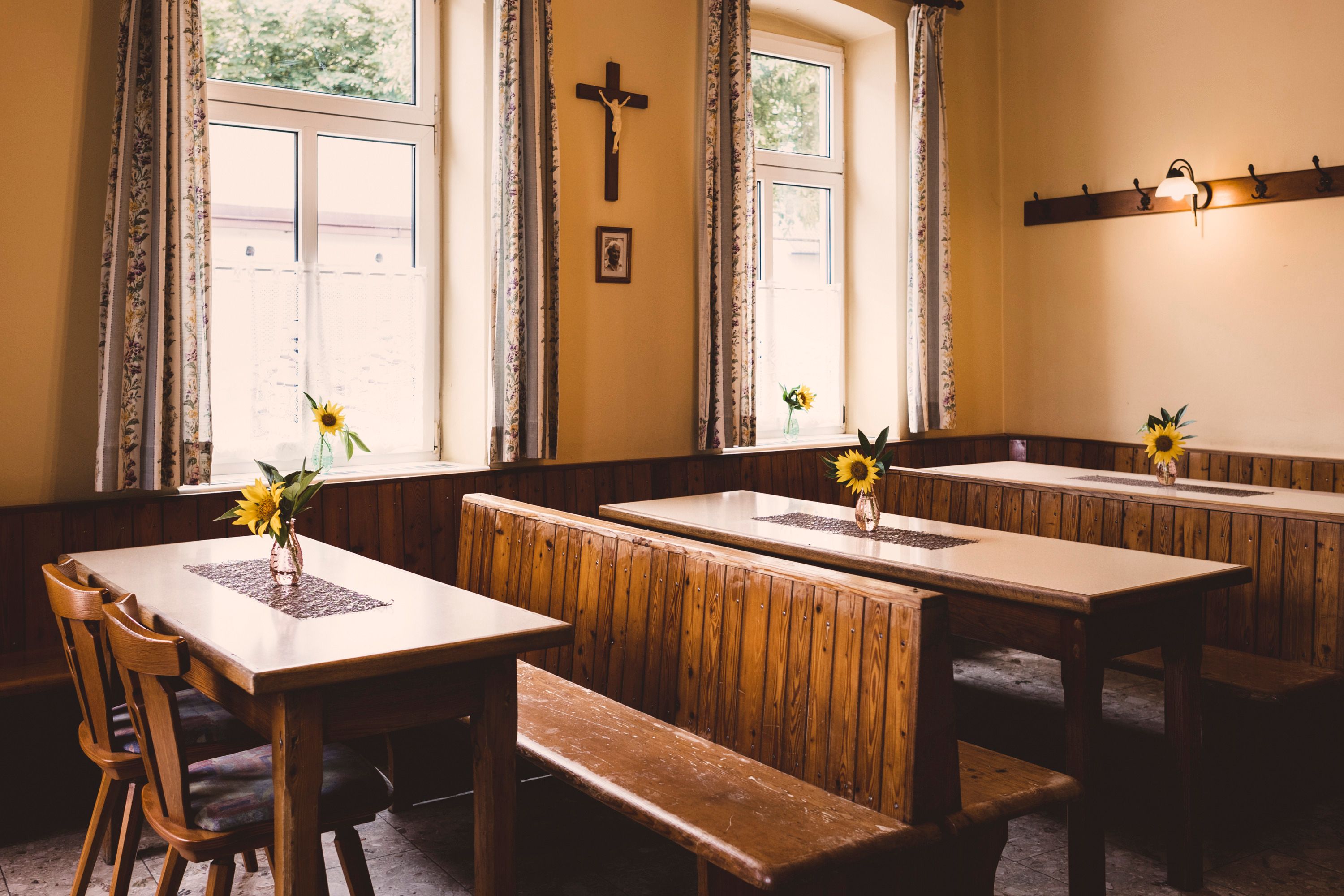 Ein rustikaler Raum mit Holzbänken und Tischen, dekoriert mit Sonnenblumen in Vasen. Ein Kreuz hängt an der Wand.