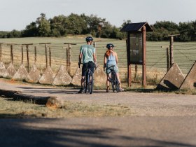 Zwei Radfahrer stehen vor einem historischen Grenzzaun mit Informationstafel.