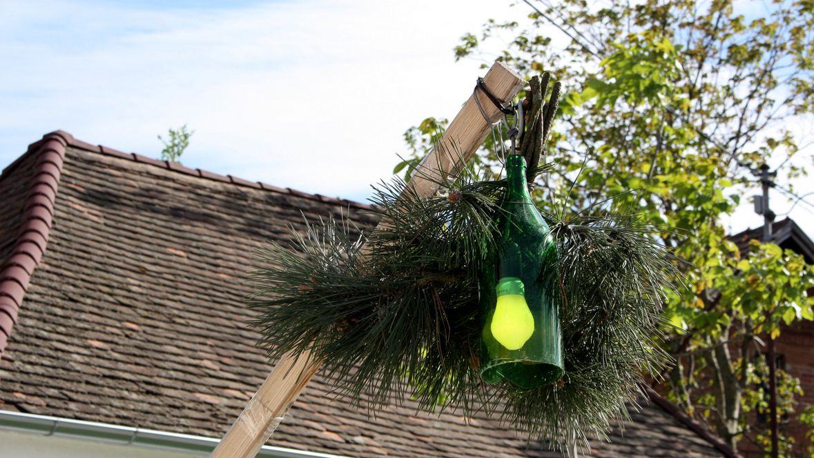 Eine grüne Flasche mit einer gelben Glühbirne hängt an einem Holzstab, umgeben von Tannenzweigen, vor einem Dach und Bäumen.