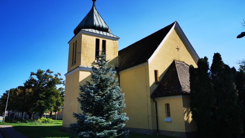 Gelbe Kapelle mit Turm und Kreuz, umgeben von Bäumen, unter blauem Himmel.