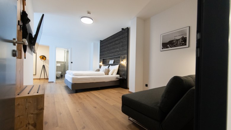 Modern eingerichtetes Hotelzimmer mit Doppelbett, Sofa und Flachbildfernseher. Helle Holzböden und ein Badezimmer im Hintergrund.
