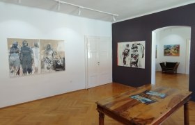 Innenansicht einer Kunstgalerie mit Holzboden, Gemälden an den Wänden und einem Holztisch in der Mitte.