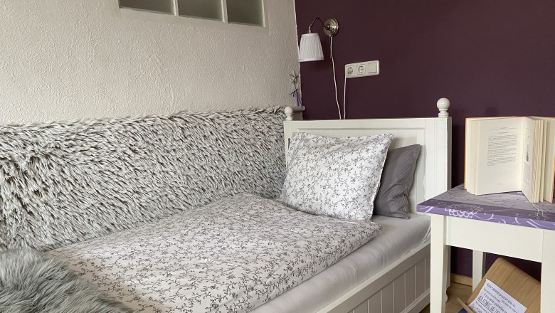 Gemütliches Schlafzimmer mit Einzelbett, lila Wand, Nachttisch mit Buch und Lampe.