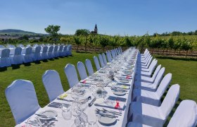 Lange, festlich gedeckte Tafel im Freien mit weißen Stühlen, umgeben von Weinbergen und einem Kirchturm im Hintergrund.