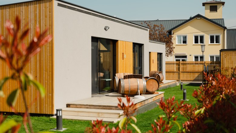 Moderne Appartements mit Holzverkleidung und Weinfässern auf der Terrasse, umgeben von grünem Rasen und einem Zaun.