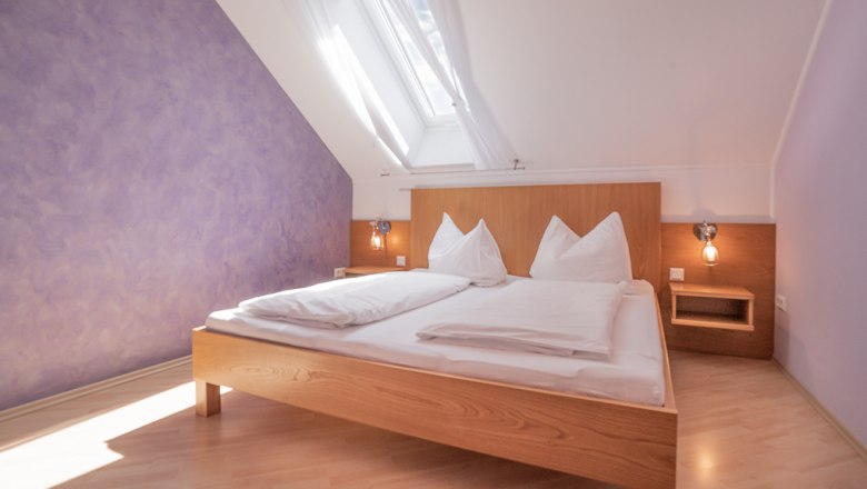 Helles Schlafzimmer mit Dachfenster, lila Wand und Holzbett.