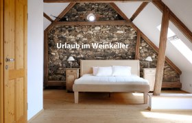 Schlafzimmer mit Holzbalken und Steinwand, Bett in der Mitte, Text 'Urlaub im Weinkeller'.