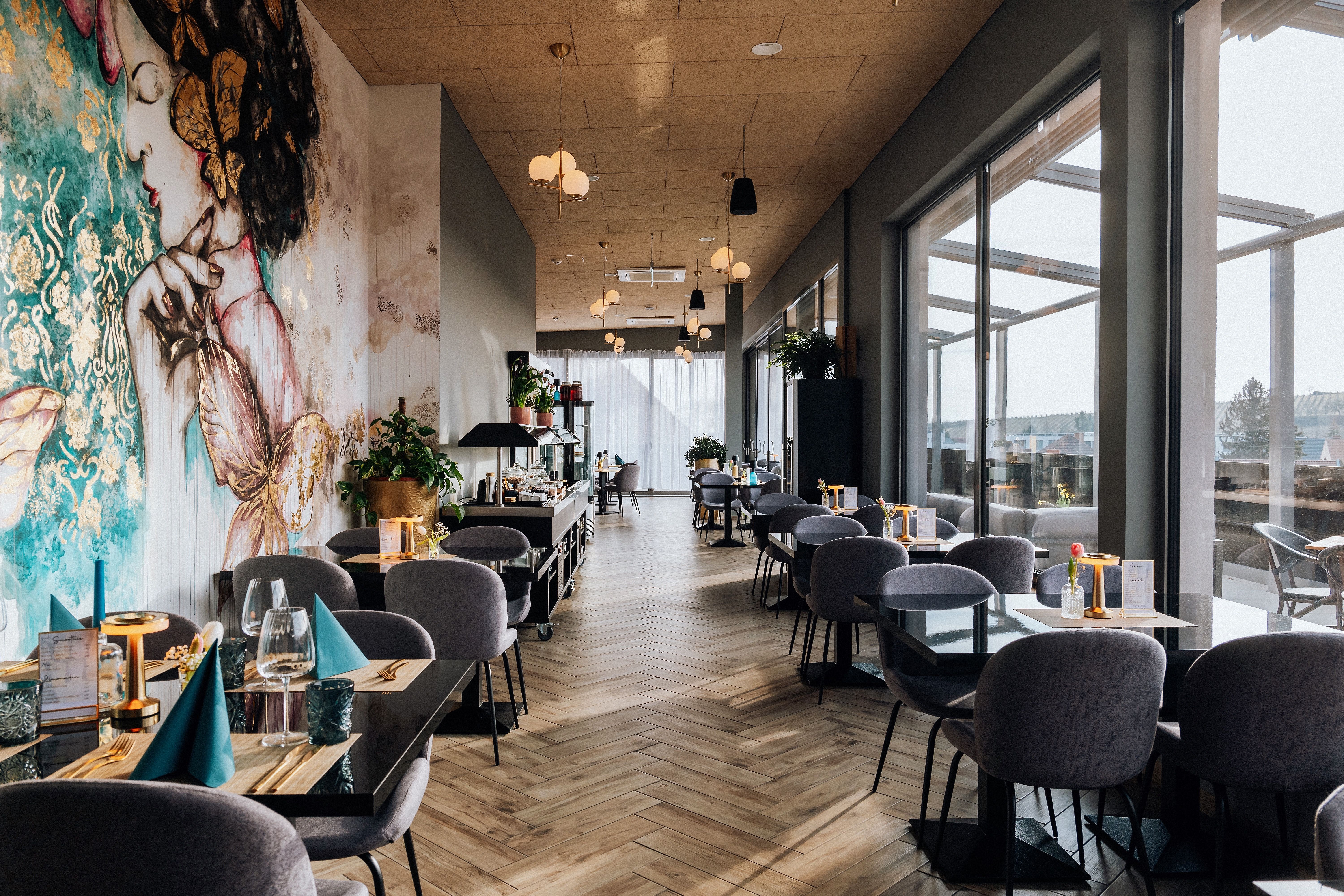 Innenansicht eines stilvollen Restaurants mit großen Fenstern und künstlerischer Wandmalerei.