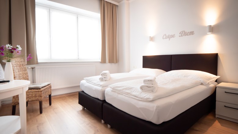 Doppelzimmer, © Doris Eminger Modernes Doppelzimmer mit zwei Betten, weißen Bettdecken und Handtüchern. Schriftzug 'Carpe Diem' an der Wand, helle Vorhänge und Holzboden.