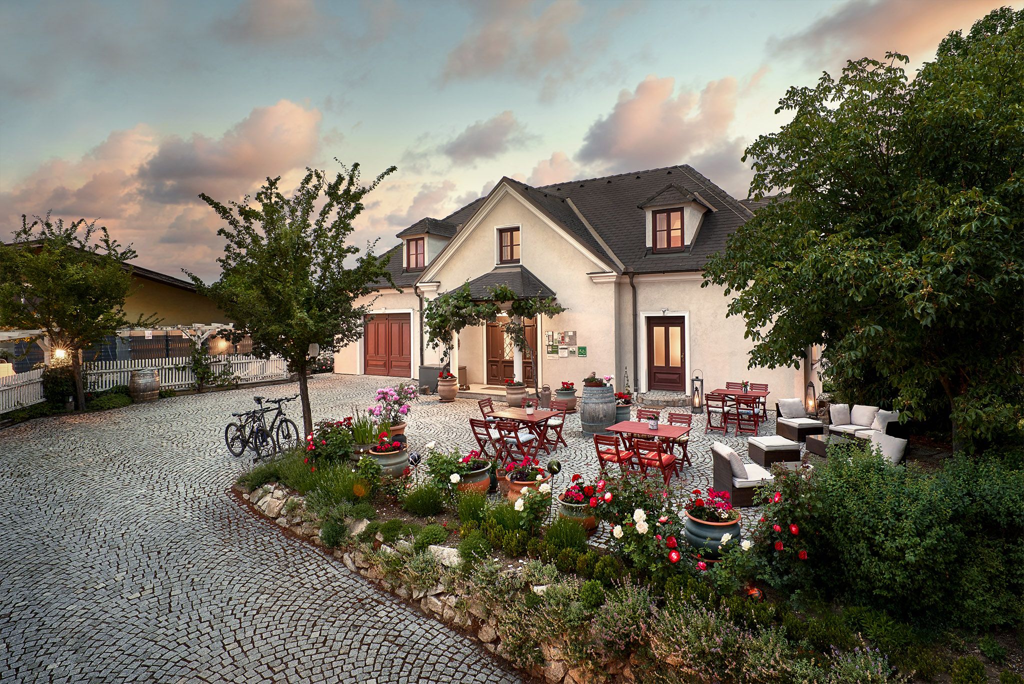 A cozy courtyard with tables, chairs and flower beds in front of a large house at sunset.