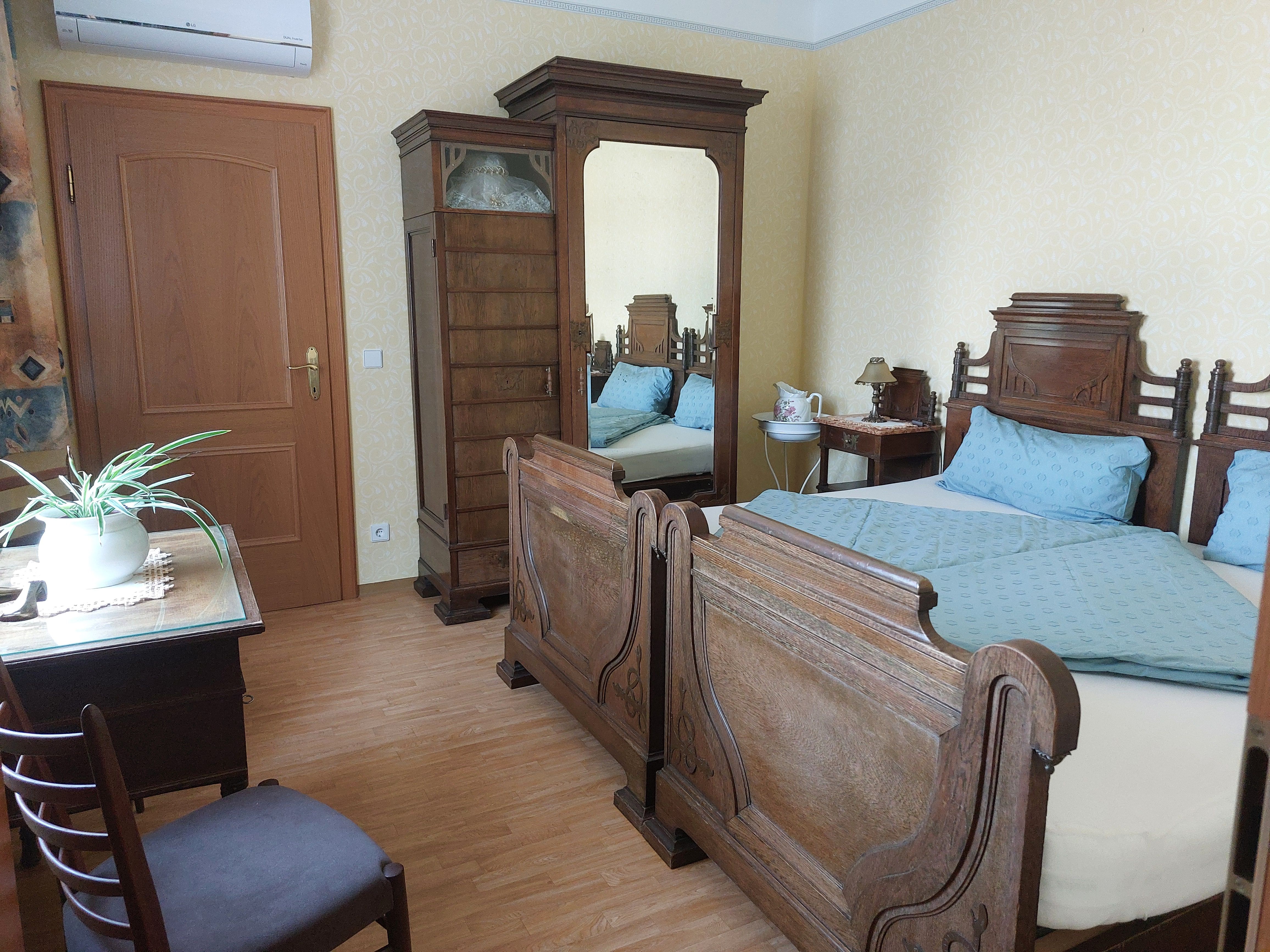 Schlafzimmer mit antiken Holzmöbeln, Doppelbett, Schrank mit Spiegel, Tisch mit Pflanze und Stuhl.