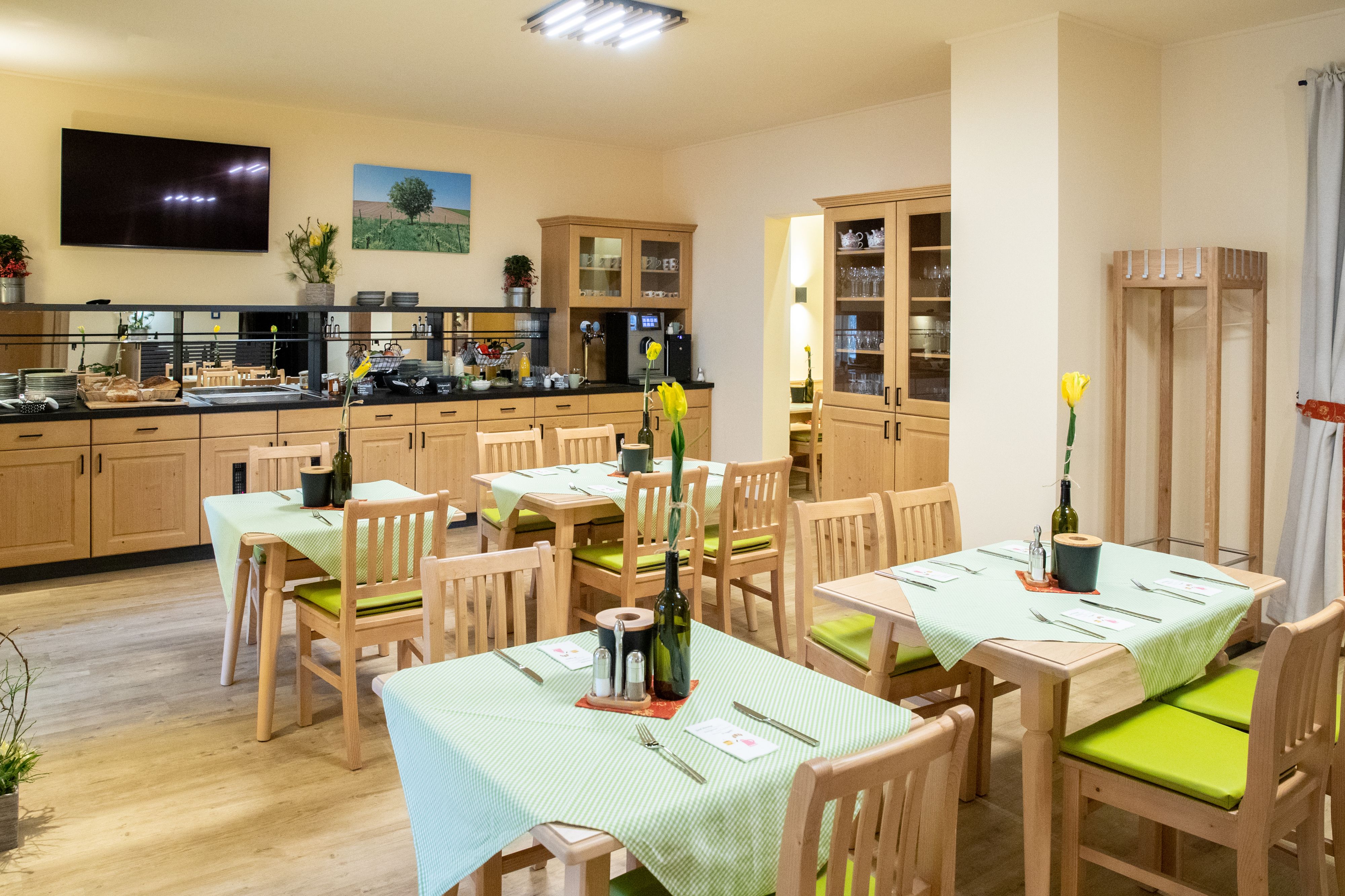Ein gemütlicher Frühstücksraum mit Holzmöbeln, gedeckten Tischen und einem Buffetbereich. An den Wänden hängen Bilder und Pflanzen dekorieren den Raum.