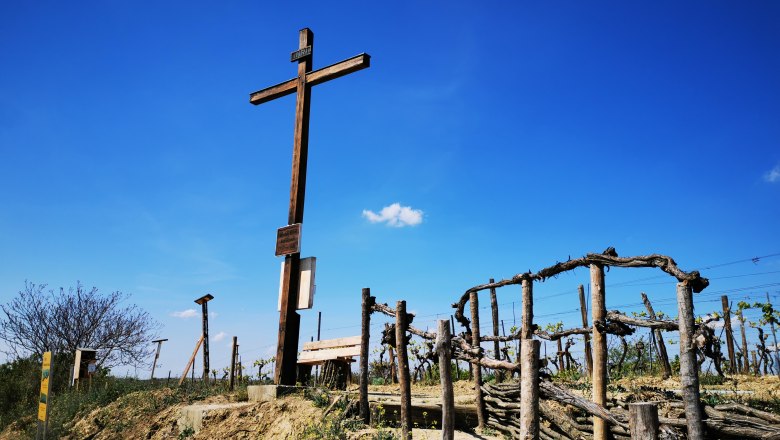 Holzkreuz auf einem H&uuml;gel mit Weinreben und blauem Himmel im Hintergrund.
