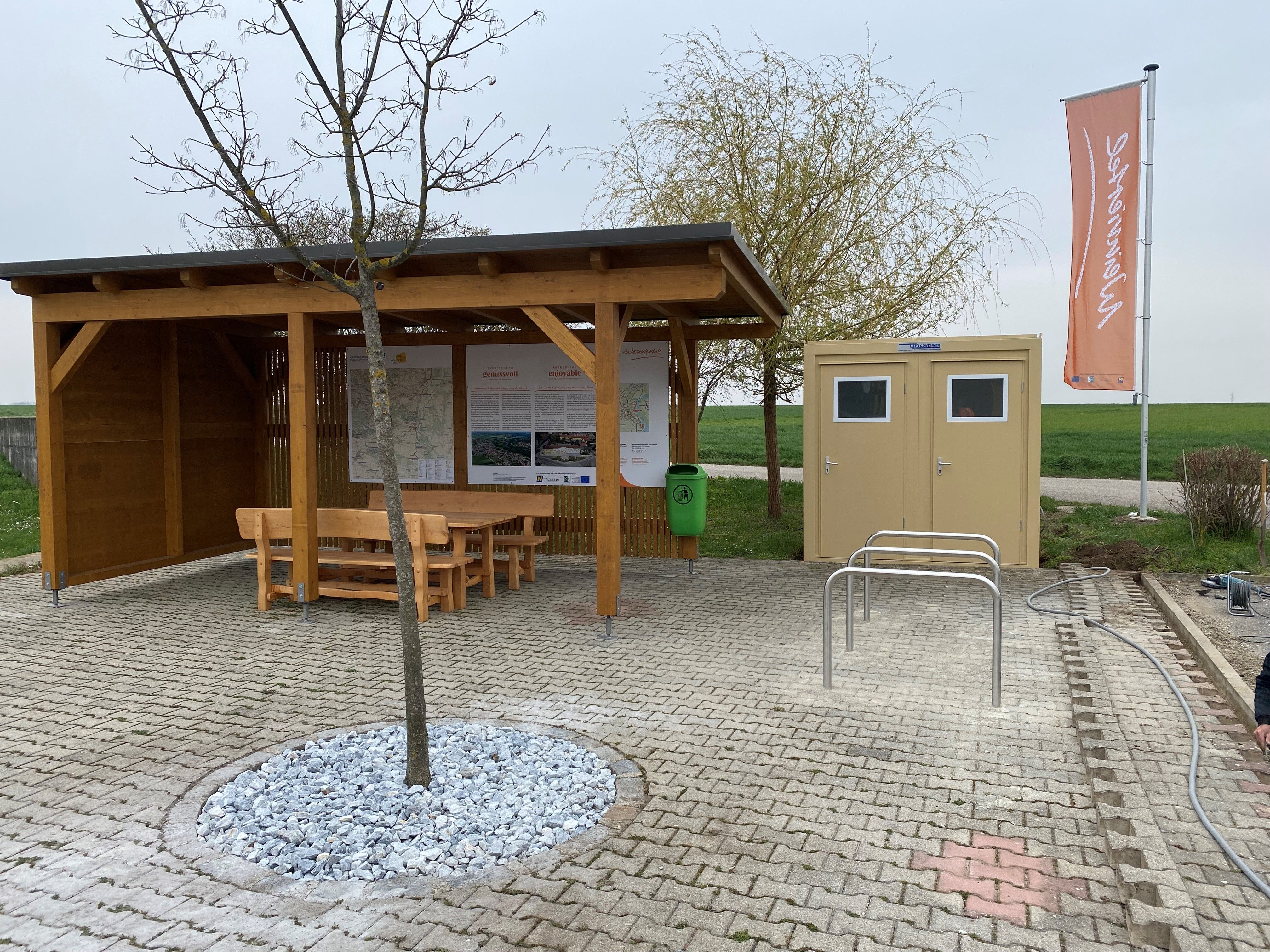 Odpočívadlo s dřevěnou chatkou, lavičkami, stojany na kola a toaletami na dlážděném prostranství.