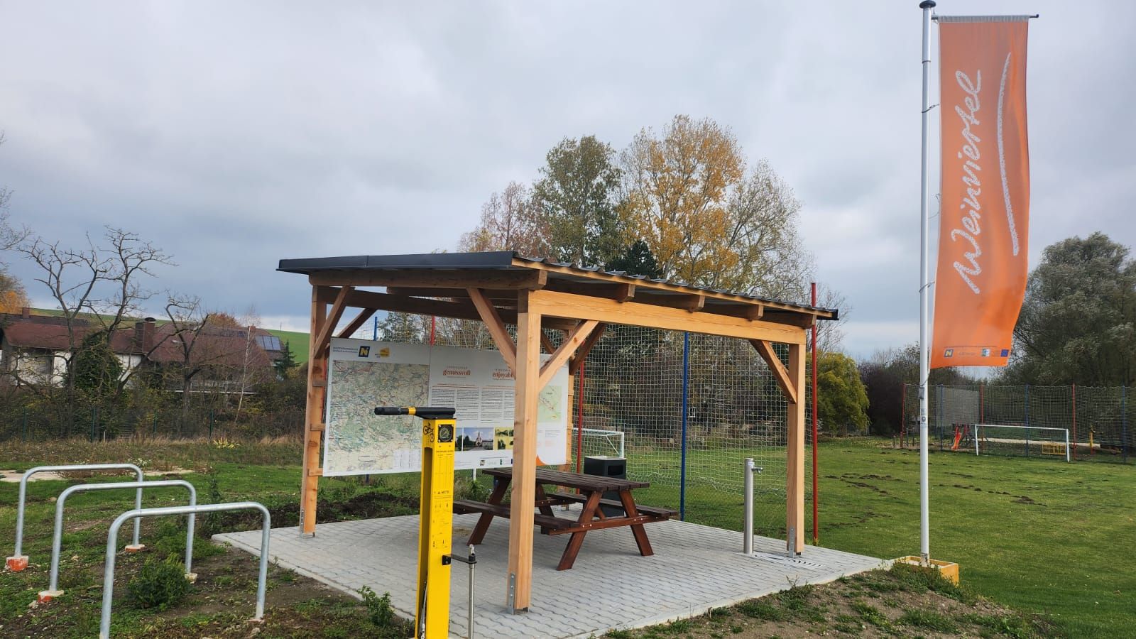 Ein Rastplatz mit Holzdach, Tisch und Bank, umgeben von Bäumen. Eine orange Fahne mit der Aufschrift 'Weinviertel' weht im Wind.