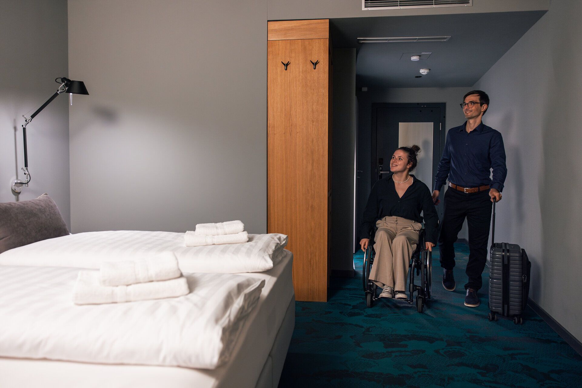 Eine junge Frau im Rollstuhl und ein Mann mit Koffer betreten ein Hotelzimmer mit Doppelbett und dunklem Teppichboden.
