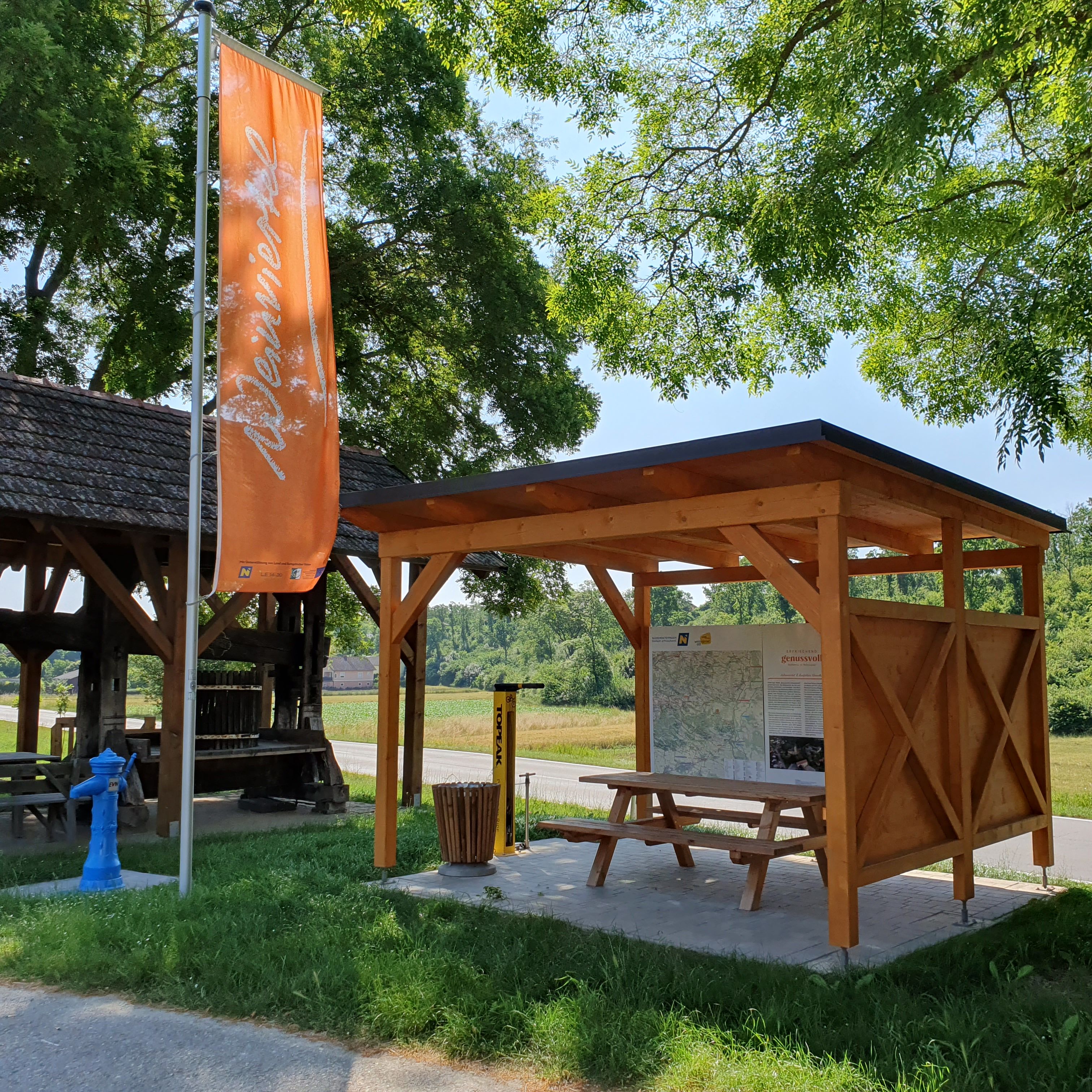 A rest area with a wooden roof and a bench, next to which is an orange flag bearing the inscription “Weinviertel.”
