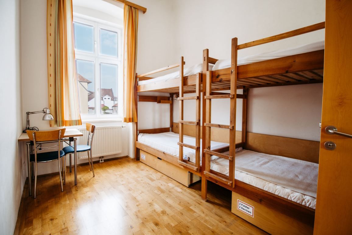 Jednoduchá izba s poschodovou posteľou, stolom a stoličkou, svetlé závesy na okne.