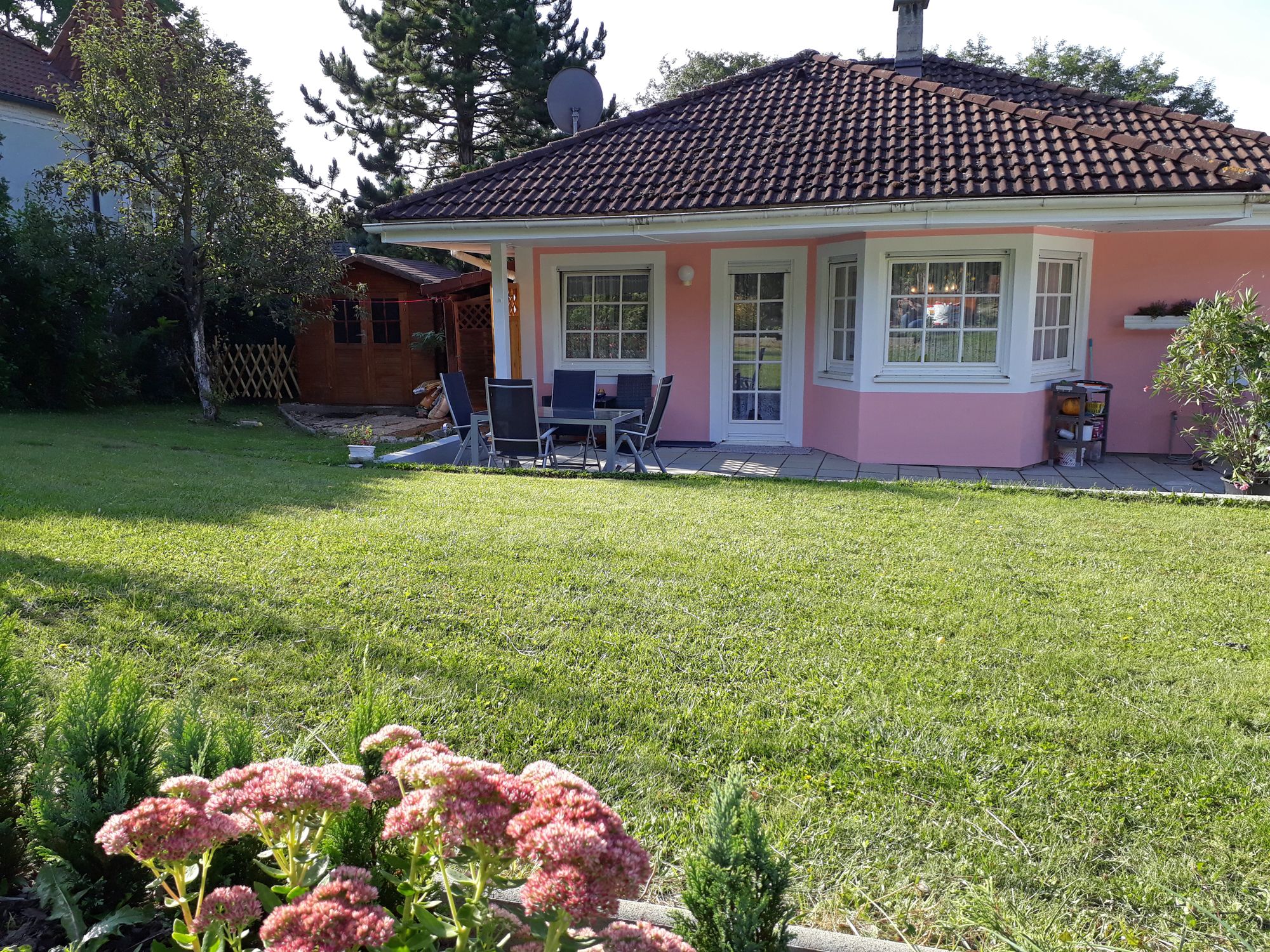 Ein rosa Haus mit Garten, Terrasse und Gartenmöbeln, umgeben von Bäumen und Blumen.