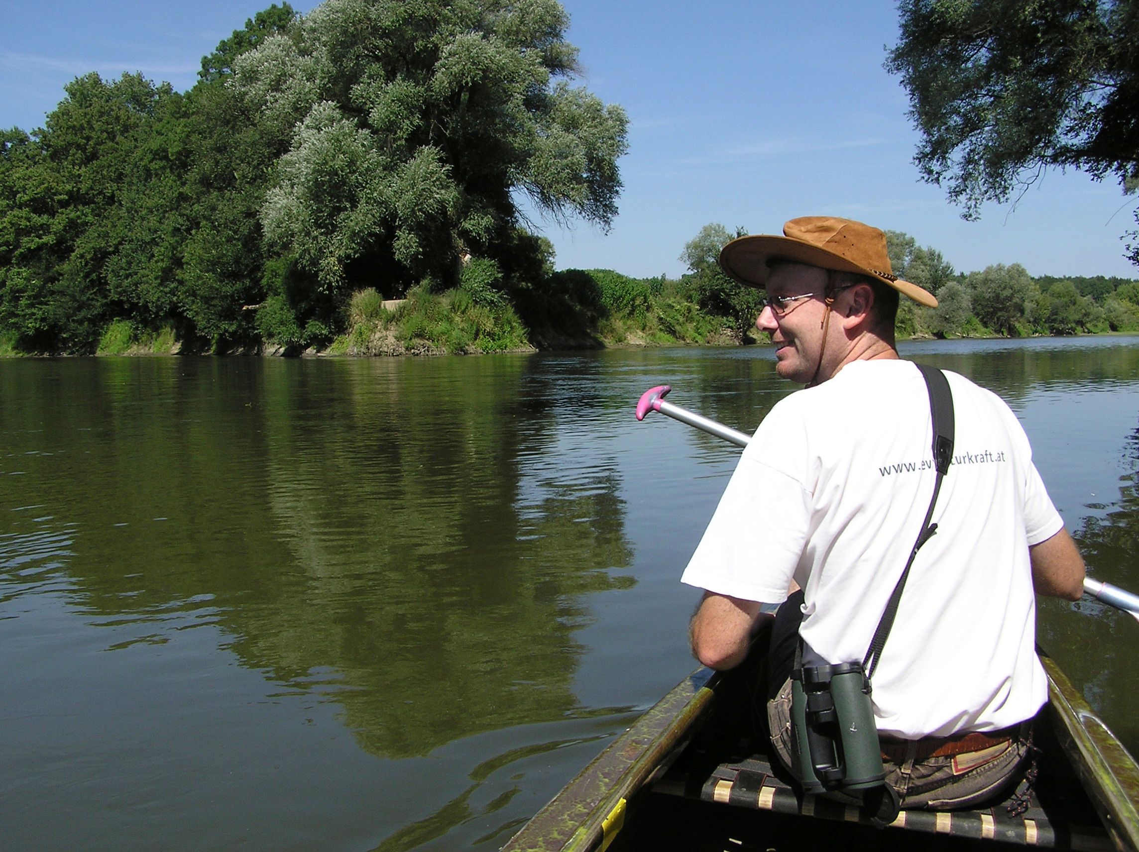 Ein Mann mit Hut paddelt in einem Kanu auf einem Fluss, umgeben von grünen Bäumen.