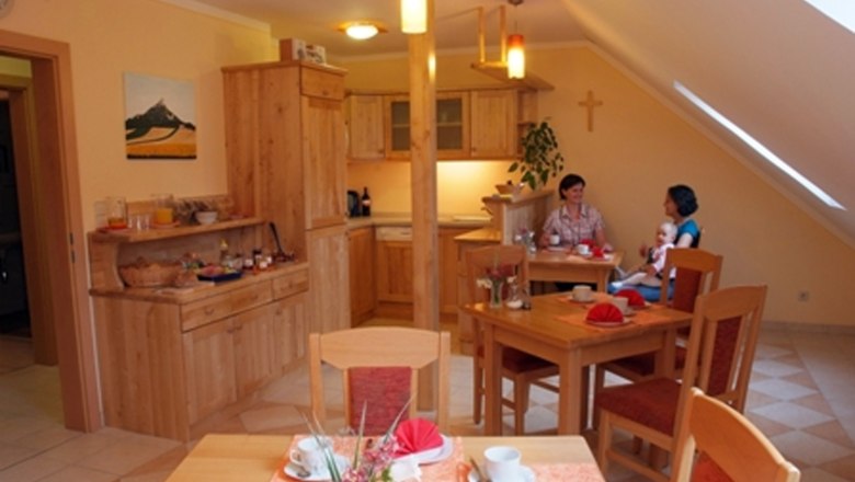 Ein gemütlicher Frühstücksraum mit Holzmöbeln, gedeckten Tischen und zwei Personen, die sich unterhalten. Ein Kind sitzt auf dem Schoß einer Person.