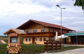 Ein traditionelles Gasthaus mit Holzbalkon und Blumendekoration, daneben ein Schild mit der Aufschrift 'Buschenschank Fam. Böswirth'.