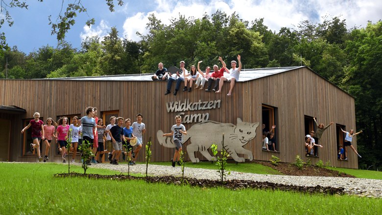 Wildkatzen Camp, © Nationalpark Thayatal / C.Milek Eine Gruppe von Kindern und Erwachsenen sitzt auf einem Holzgebäude mit der Aufschrift 'Wildkatzen Camp'. Einige Kinder stehen und springen davor.