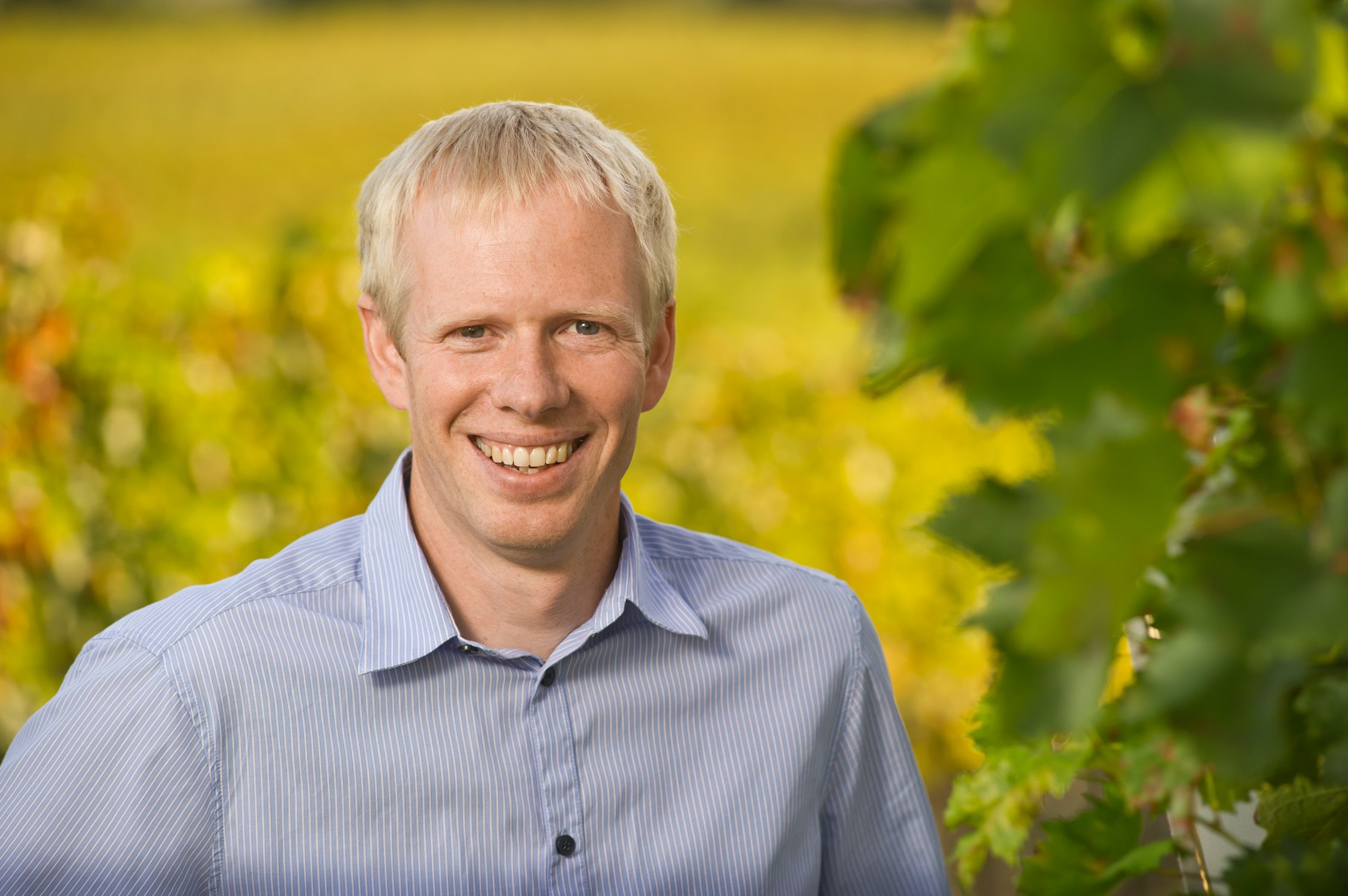 Ein lächelnder Mann in einem blauen Hemd steht vor einem unscharfen Hintergrund mit grünen Blättern.
