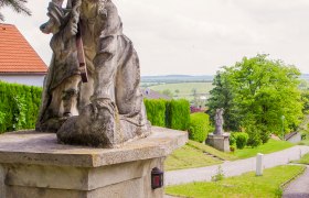 Steinstatue auf einem Sockel mit grüner Landschaft im Hintergrund.