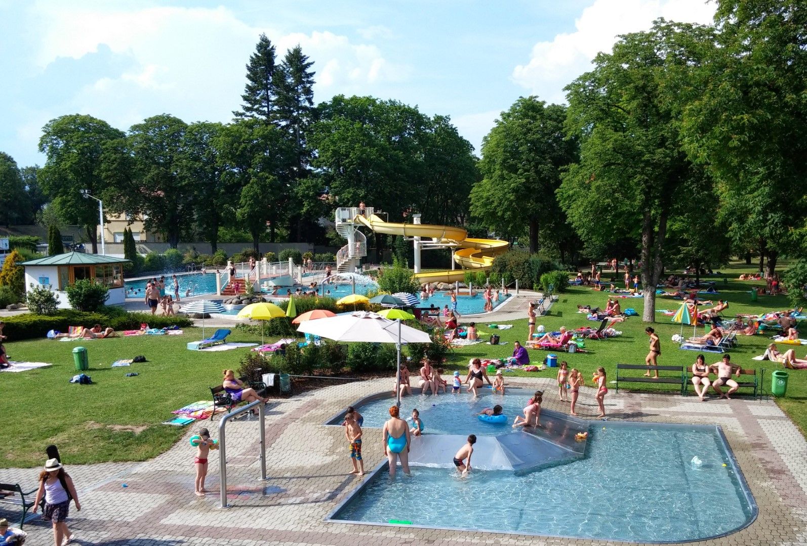 Ein Freibad mit mehreren Pools, einer gelben Wasserrutsche und vielen Menschen, die auf der Wiese entspannen.
