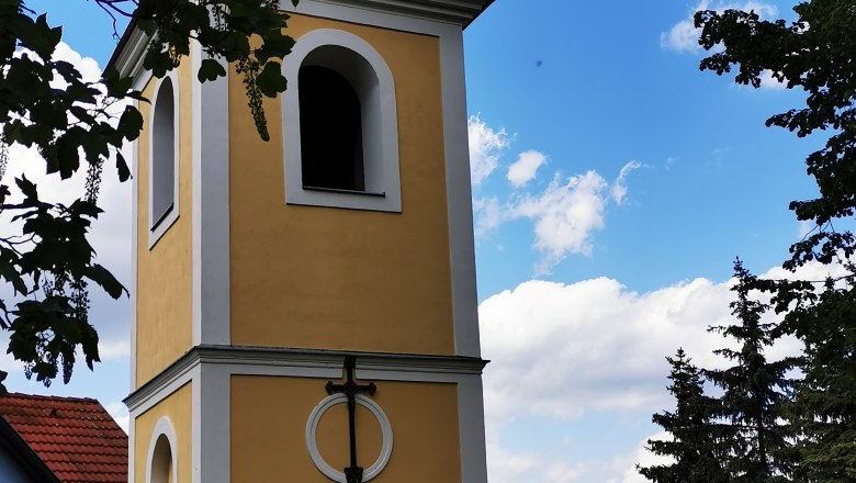 Glockenturm in Ragelsdorf vor blauem Himmel und Bäumen.