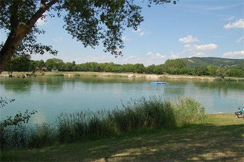 Ein ruhiger See mit Schilf im Vordergrund und bewaldeten H&uuml;geln im Hintergrund.