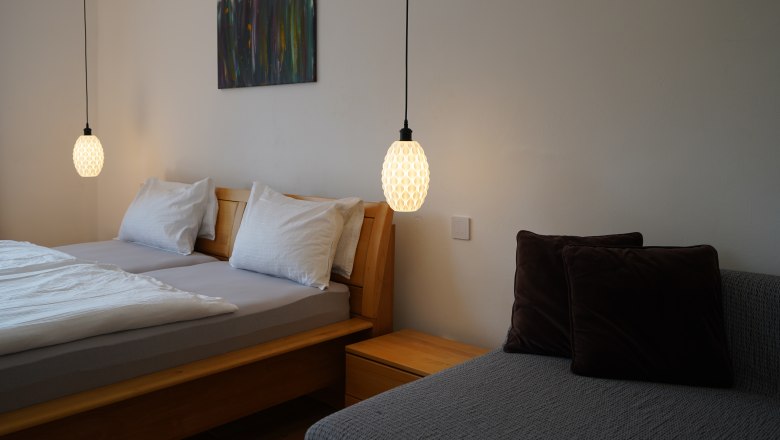 Schlafzimmer mit Doppelbett, zwei H&auml;ngelampen und einem Sofa mit Kissen.