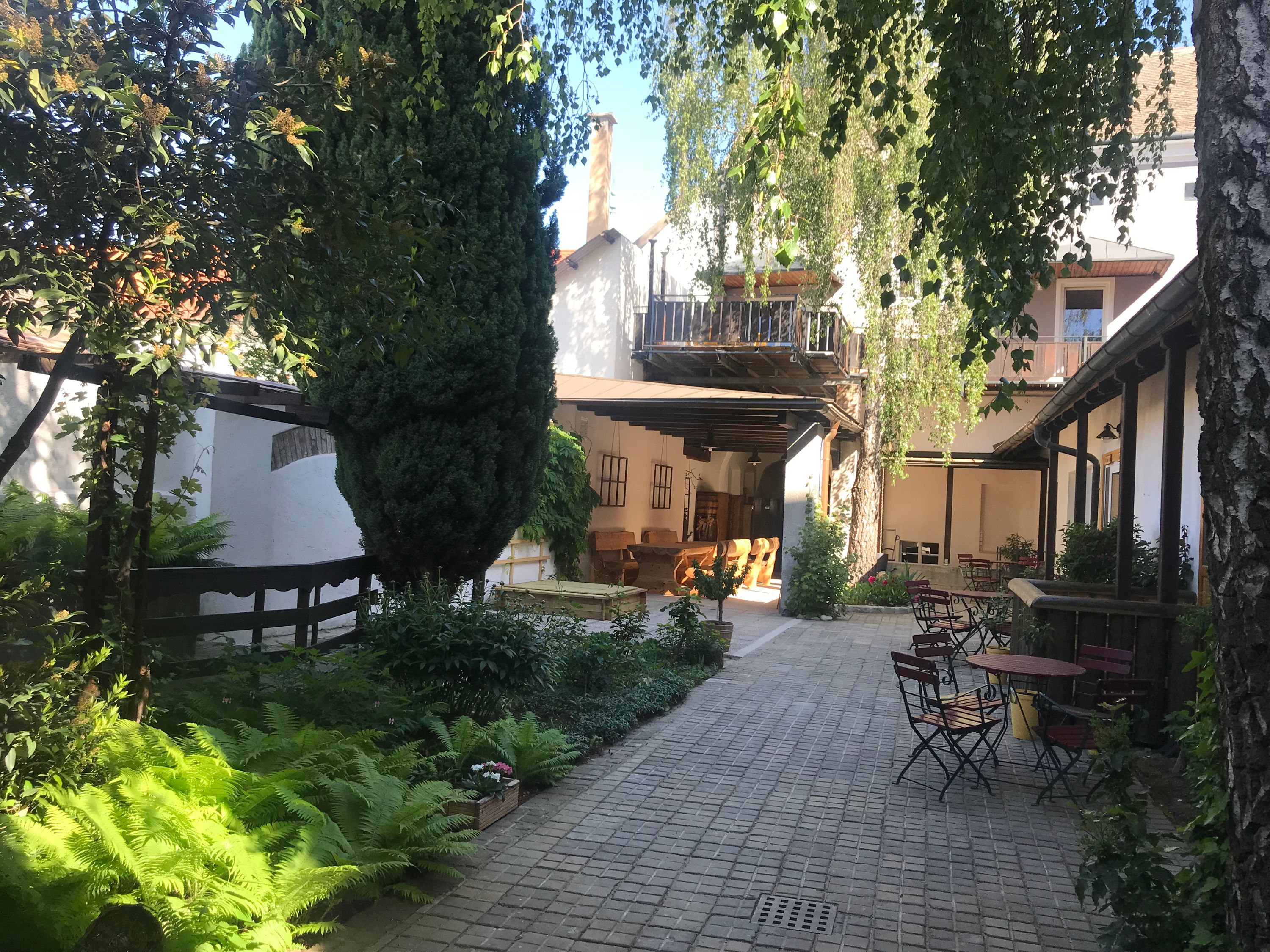 Ein ruhiger Innenhof mit Bäumen, Pflanzen und Gartenmöbeln unter blauem Himmel.