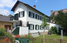Ein wei&szlig;es Geb&auml;ude mit gr&uuml;nen Fensterl&auml;den, umgeben von einem Garten, mit einer Burg im Hintergrund.