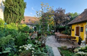 Ein gemütlicher Garten mit gelbem Gebäude, Holzbänken und üppiger Vegetation unter blauem Himmel.