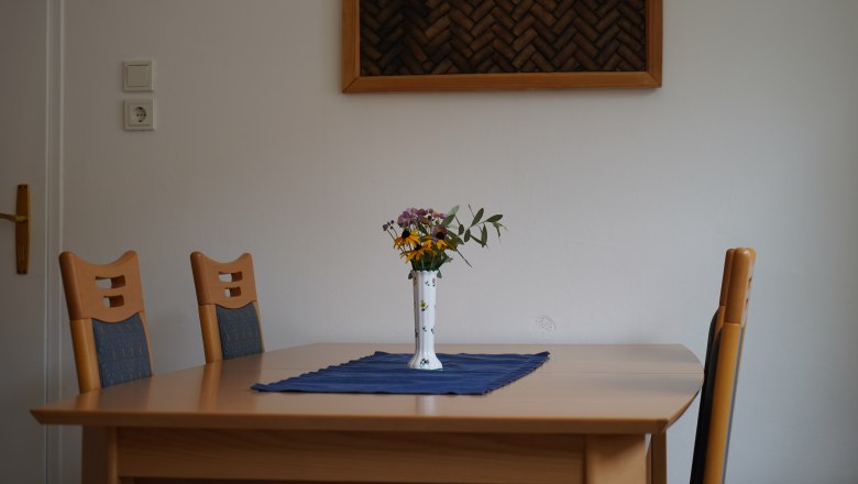 Ein Tisch mit vier St&uuml;hlen, einer Vase mit Blumen und einem blauen Tuch in einem Raum mit wei&szlig;er Wand und einem Wanddekor.