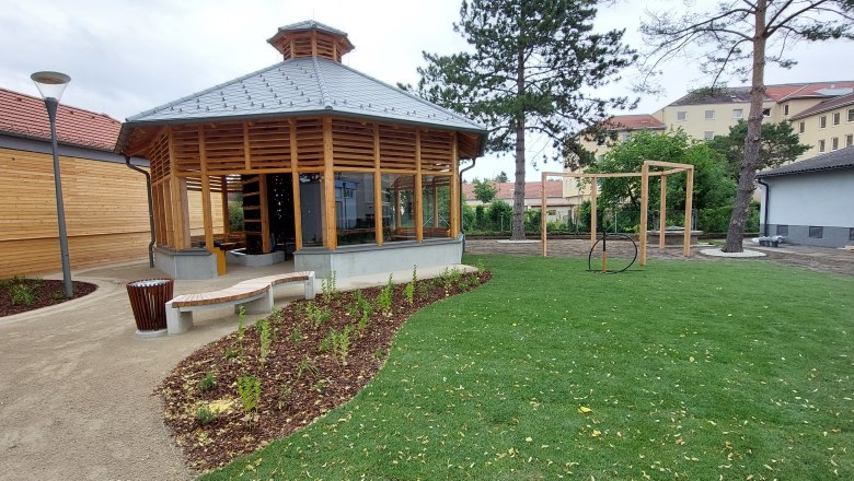 Ein modernes Inhalatorium mit Holzkonstruktion und Glasfenstern in einem gepflegten Garten mit Rasen und Bäumen.