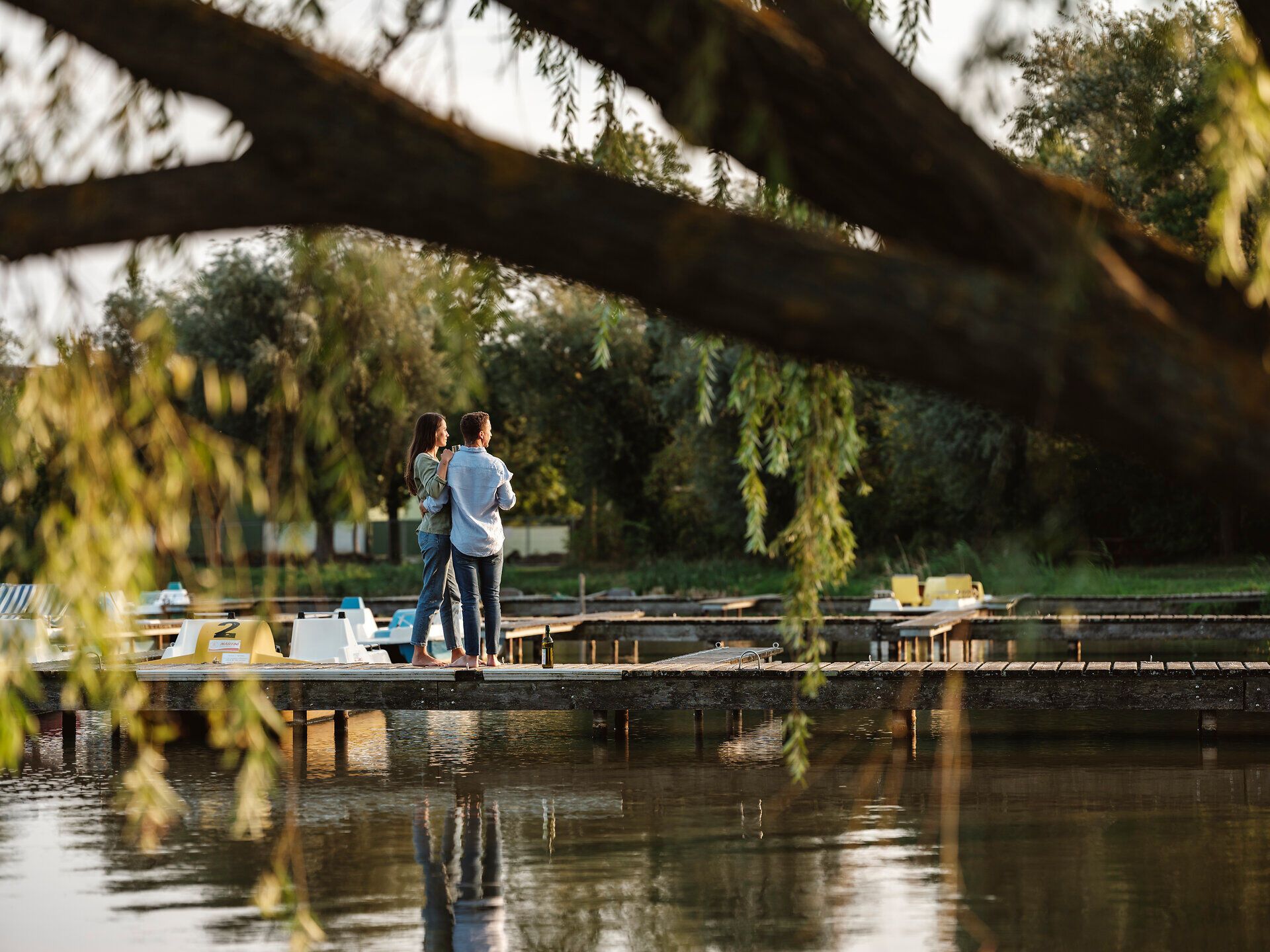 Ein romantischer Moment am Steg, wo die sanften Wellen des Sees im Licht der Abendsonne glitzern. Die umgebenden Weidenbäume verleihen der Szenerie eine friedliche Atmosphäre, ideal für eine wohlverdiente Pause mit einem Glas Wein.