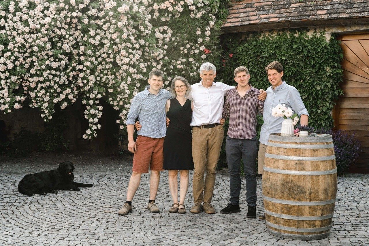 Eine Familie steht lächelnd vor einer blühenden Wand, mit einem Hund im Hintergrund und einem Weinfass mit Blumen rechts.