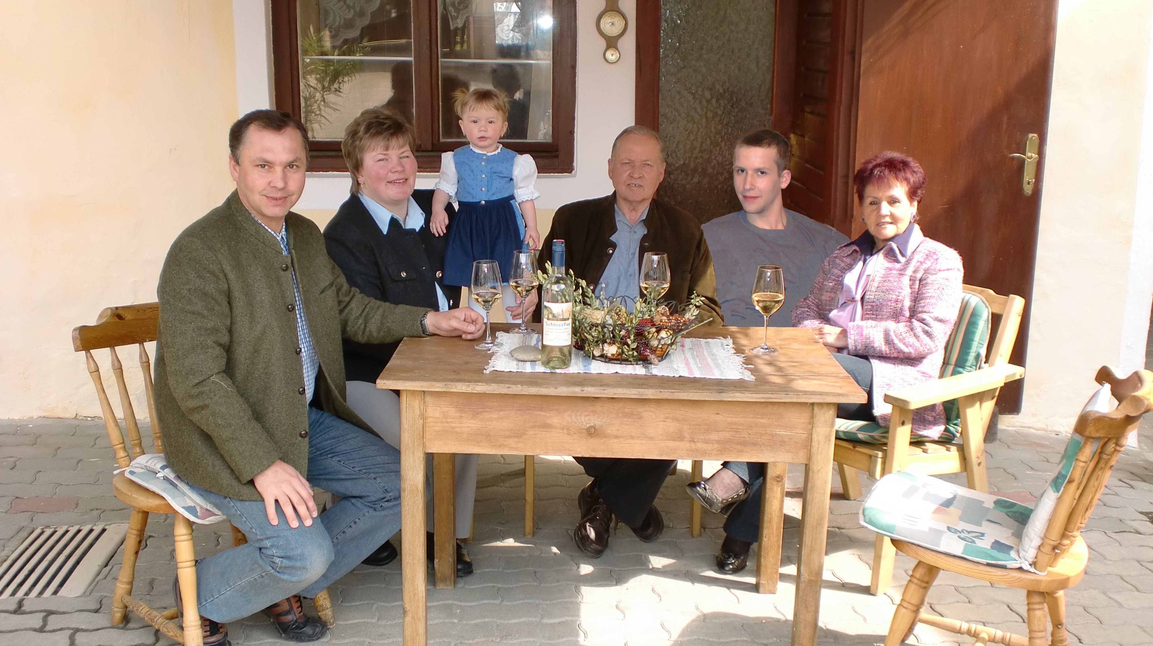 A family is sitting at a wooden table outside drinking wine.