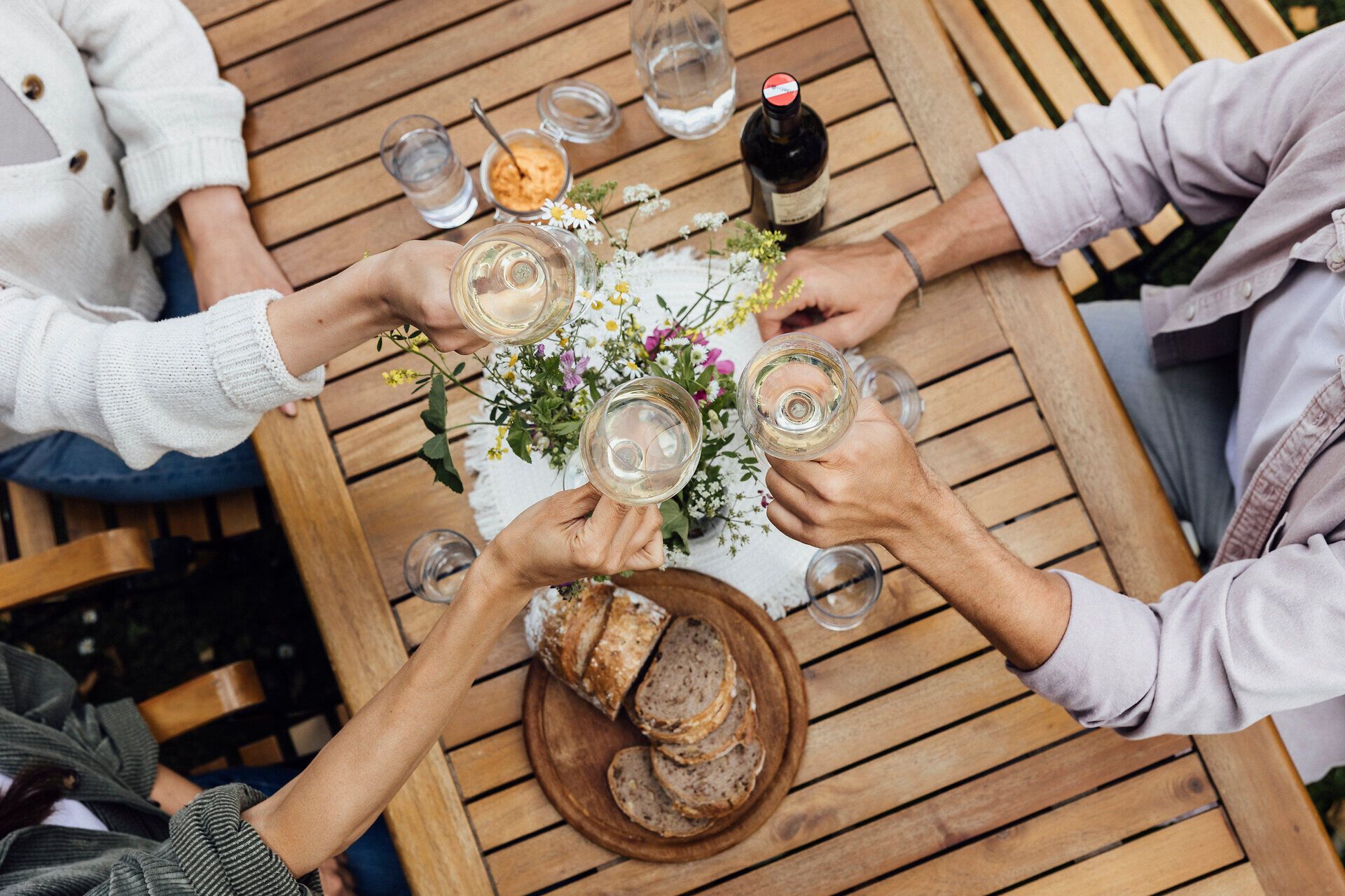 Inmitten einer idyllischen Atmosphäre stoßen Freunde mit Gläsern auf die schönen Momente des Lebens an. Der Tisch ist liebevoll gedeckt mit frischem Brot und einer bunten Auswahl an Köstlichkeiten, während die Blumen in der Mitte des Tisches die Freude des Augenblicks unterstreichen.