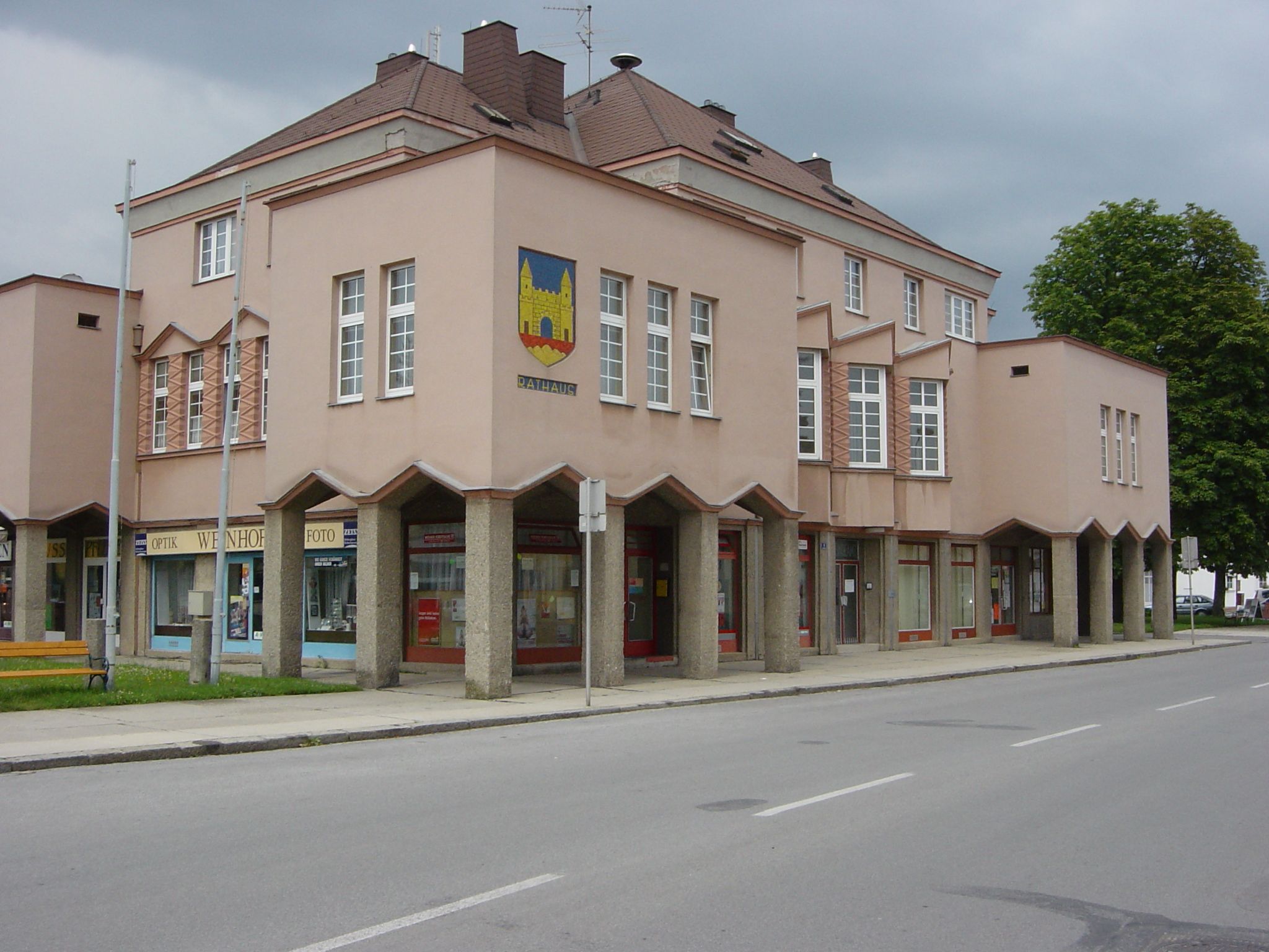 Ein modernes Rathausgebäude mit rosa Fassade und großen Fenstern. Es hat ein Wappen an der Ecke und Geschäfte im Erdgeschoss.